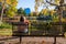 Little lonely girl with a blue balloon and a bunch of a fall leaves sits on an empty bench in a fall park.