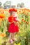 Little longhaired blonde girl in red dress at field of poppies with wreath at her head on summer sunset