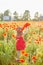 Little longhaired blonde girl in red dress at field of poppies with wreath at her head on summer sunset