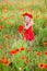 Little longhaired blonde girl in red dress at field of poppies with wreath at her head on summer sunset