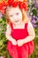 Little longhaired blonde girl in red dress at field of poppies with wreath at her head on summer sunset