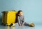 Little male in gray overall, yellow sunglasses, rubber boots. Sitting on floor near suitcase, smiling, posing on blue background