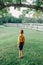 Little preschool Caucasian boy in yellow shirt and jeans shorts with suspenders standing alone in park country-side village