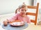 Little preschool girl, finishing her meal, sitting at the table on a chair near an empty plate holding a fork, finished meal