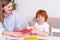 Little red-haired boy  with a nanny or mother or teacher  sit at the table in the room and cuts out of colored paper with scissors