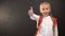 Little schoolgirl showing thumbs-up on camera against blackboard, education