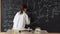 A little scientist girl in a white coat writes a formula on a blackboard with chalk. Research and education at school.
