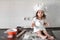 Little smiling baby girl baker in white cook hat and apron kneads a dough on tle kitchen