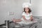 Little smiling baby girl baker in white cook hat and apron kneads a dough on tle kitchen