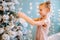 Little smiling blonde girl decorating christmas tree with toys and glass balls at home. Fir-tree is coverd with white