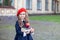 Little smiling girl in a red beret holds a stack of books with an apple near school building. Cute schoolgirl of primary school on