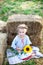 Little smiling girl sits on a roll of haystack in garden and holds a sunflower. A child sits on a straw and enjoys nature in count