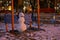 A little snowman sits on a swing on a blurry background of the evening courtyard and apartment buildings.