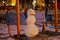 A little snowman sits on a swing on a blurry background of the evening courtyard and apartment buildings.