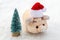 Little souvenir Christmas tree next to a toy pink pig in a red Santa Claus hat on white snow. Concept on the theme of celebrating