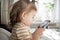 Little toddle girl playing with smartphone laying on the bed at home. Caucasian toddler girl.