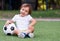 Little toddler boy sitting with legs crossed on football field in summer day with soccer ball. Happy active child