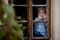 Little toddler child, boy with blue shirt, holding single daisy flower, sitting behind old window, looking outdoors
