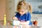 Little toddler girl making craft lantern with paper cups, colorful pompoms and glue during pandemic coronavirus