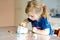 Little toddler girl making craft lantern with paper cups, colorful pompoms and glue during pandemic coronavirus