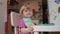 Little two years old blond blue-eyed girl grimaces while having her dining, 4K shot