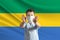 Little white boy in a protective mask on the background of the flag of Gabon. Makes a stop sign with his hands, stay at