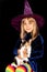 Little witch in a huge hat on her head on a black background. The girl is holding a three-haired cat. Halloween costume