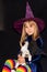 Little witch in a huge hat on her head on a black background. The girl is holding a three-haired cat. Halloween costume