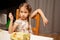 Little young girl eating her food holding a fork in her hand sitting on a chair in a sloppy, careless relaxed pose / manner