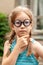 Little young school age girl in big funny quirky glasses thinking, wondering, deep in thought. Outdoors portrait, one person face