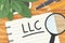 LLC (Limited Liability Company) written on spiral notebook with magnifying glass on wooden desk, flat lay view