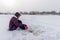 A lone angler girl is concentrating on catching fish with a winter fishing rod sitting on a substrate on the ice among flakes