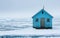 A lone cabin stands amidst a serene, icy landscape, surrounded by floating ice and reflected in the still water.