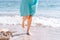 A lone girl in a blue dress walks along the waterline of a sandy beach. Women`s feet along the beach. The concept of travel,