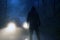 A lone hooded figure standing in front of car headlights in the middle of a country road on a spooky, eerie, moody foggy night