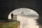 Lone woman silhouette walking under old stone arched bridge on canal walk along tow path footpath into morning sunrise with frost