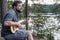 Lonely bearded man plays a melody on a guitar and is sad, sitting on a hill, against a background of trees and a lake