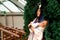 A lonely beautiful girl with multi-colored dreadlocks in a white dress is standing on a red stairs near, green cypress, thuja tree