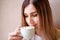 Lonely blonde girl sitting on a windowsill with a white cup with coffee with a heart, romantic mood.