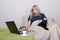 A lonely elderly woman in glasses sits on a sofa under a rug with a laptop and a black cat