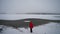 A lonely happy woman wearing a red jacket is approaching a half-frozen lake during Christmas with moody cloudy weather