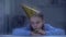 Lonely little orphan in birthday hat with party blower behind rainy window