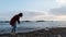 Lonely melancholy woman in casual warm wear throws stones into the water at sunset with sea and cloudy sky on background