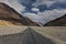 A Lonely Metal Road amidst Ladakh\\\'s Empty Valley, with Clear Blue Sky, Fluffy Clouds, and Majestic Snowy Mountains in Sight.