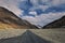A Lonely Metal Road amidst Ladakh\\\'s Empty Valley, with Clear Blue Sky, Fluffy Clouds, and Majestic Snowy Mountains in Sight.