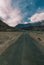 A Lonely Metal Road amidst Ladakh\\\'s Empty Valley, with Clear Blue Sky, Fluffy Clouds, and Majestic Snowy Mountains in Sight.