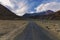 A Lonely Metal Road amidst Ladakh\\\'s Empty Valley, with Clear Blue Sky, Fluffy Clouds, and Majestic Snowy Mountains in Sight.