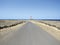 Lonely paved road in arid area towards red and white maritime lighthouse under blue ocean and sky. Lunatic coastal landscape.