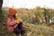 A lonely preschooler girl in an orange hat and a brown coat sits on the ground under an old big tree. autumn horizontal photo of a