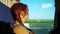 A lonely red-haired traveler girl looks out the bus window at road transport, nature and the blue sky.